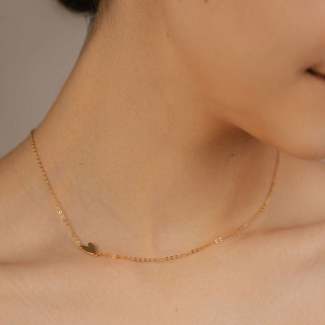 a woman is wearing a minimalist heart necklace gold