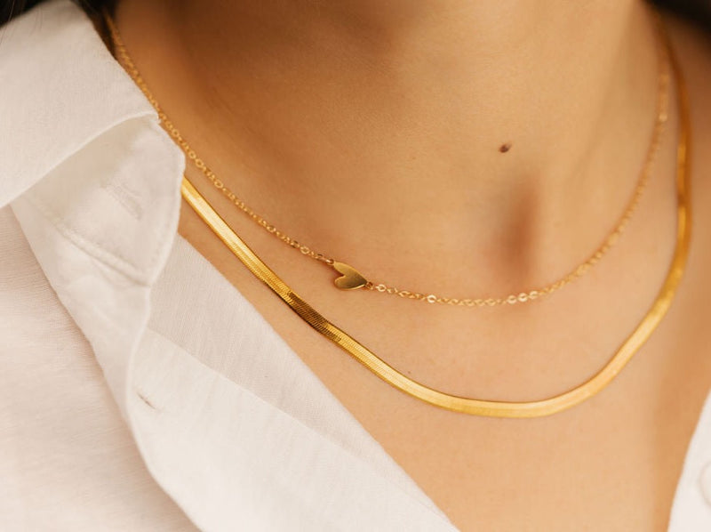 A woman is wearing two necklaces. One necklace is gold vermeil heart sideways necklace. The other one is herringbone necklace. Both made in Canada