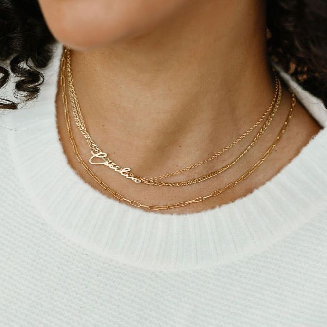 Cursive style nameplate necklace layered with gold chains.